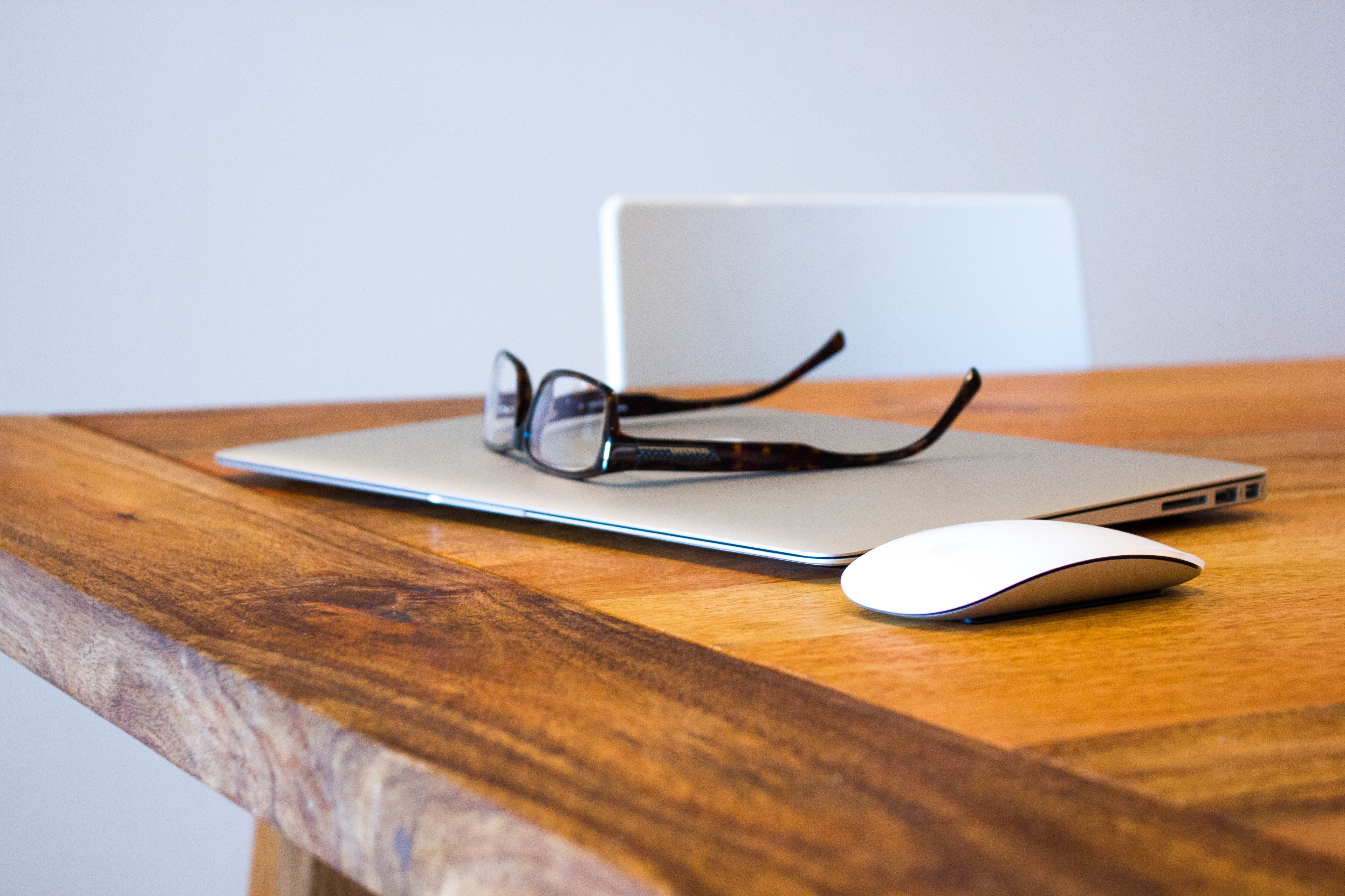 Laptop, glasses, and a slim white computer mouse are sitting in a close up shot of a wooden table, in front of a white chair and a white wall.